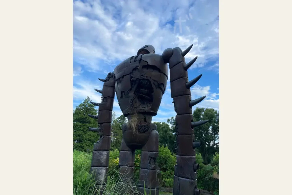 Back view of the Robot Soldier from Laputa standing on the rooftop of the Ghibli Museum, grass growing from its waist beneath a bright blue sky.