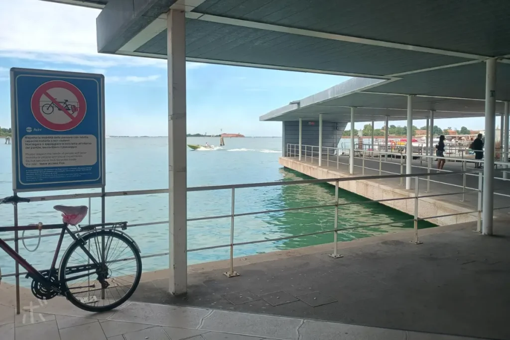ヴェネツィア・リドのフェリー乗り場で、自転車と標識の奥にラグーンの水辺が広がっている風景。