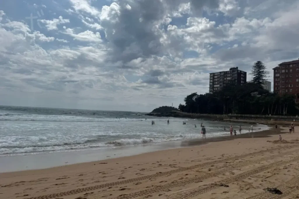 湾曲する砂浜と波打ち際、岬と建物が見えるマンリー・ビーチの景色