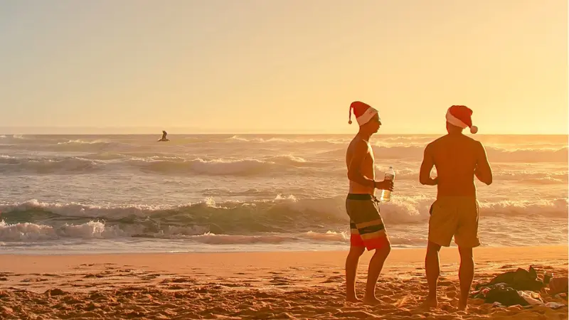 夕暮れのオーストラリアのビーチで、サンタ帽をかぶった人たちが短パン姿で過ごす真夏のクリスマス風景。