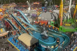 シドニー・イースター・ショーの遊園地エリアを上から見下ろした風景。ウォーターライドやアトラクション、人々の賑わいが広がるSeen Through Someone’s Eyesシリーズのアイキャッチ画像。