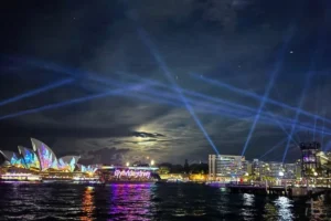 Light beams over Sydney Harbour at night with the illuminated Opera House and reflections shimmering across the water